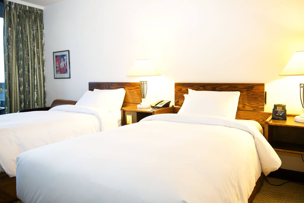 Une chambre d’hôtel avec deux lits, des têtes de lit en bois, des tables de chevet avec des lampes, un téléphone et un tableau encadré au mur.