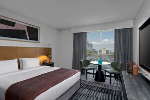 A hotel room with a large bed, a desk with a chair, a small table with two chairs, a TV, and a view of the city through large windows.