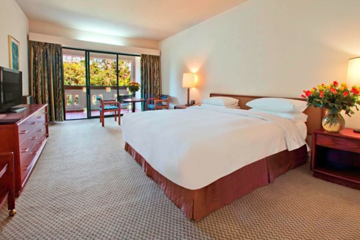 A hotel room with a large bed, a desk, a chair, a television, and a balcony with curtains.