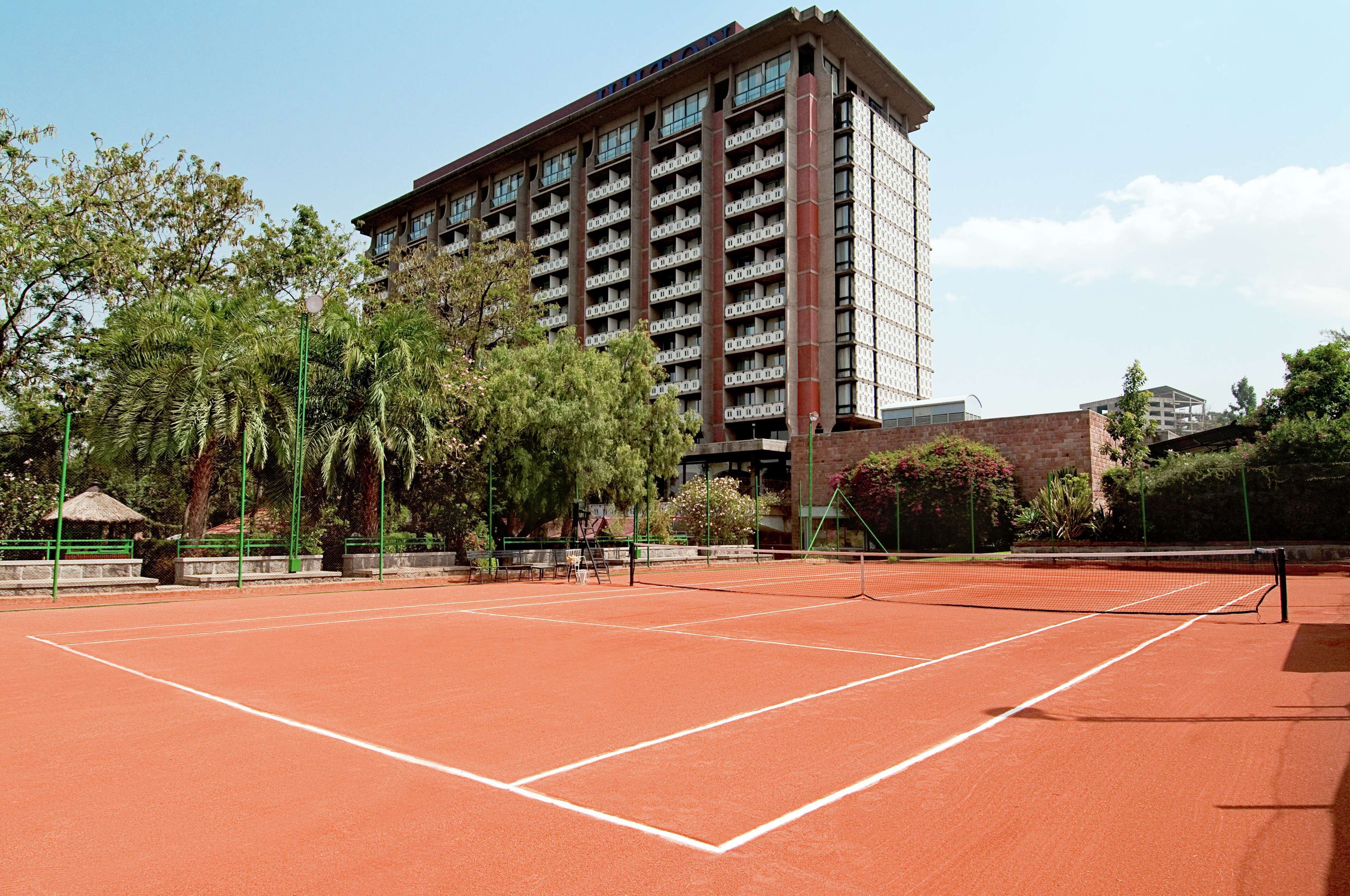 Een tennisbaan met een net, omgeven door bomen en een gebouw op de achtergrond.