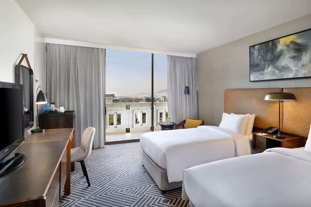 Une chambre d’hôtel avec deux lits, un bureau avec une télévision, une chaise, une table de chevet avec une lampe et une grande fenêtre donnant sur des bâtiments et des montagnes.