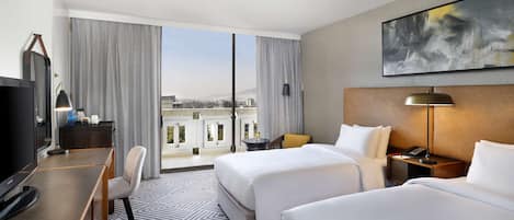 Habitación de hotel con dos camas, un escritorio con televisor, una silla, una mesita de noche con lámpara y un amplio ventanal con vistas a edificios y montañas.