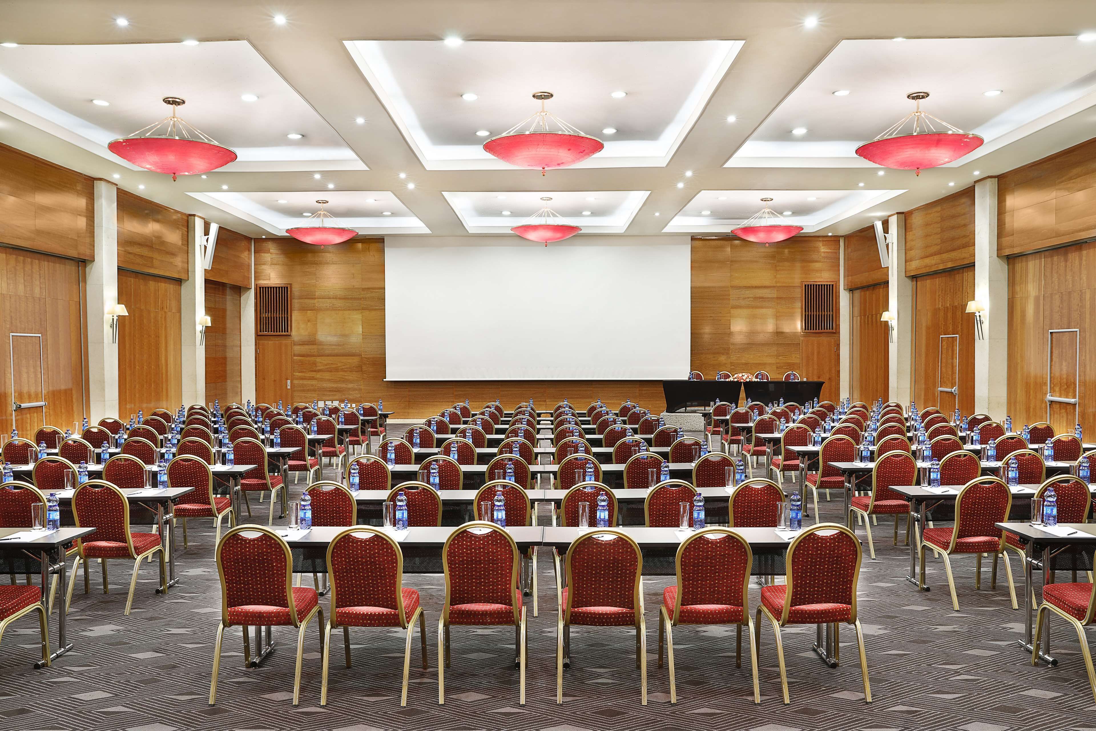 Une salle de conférence avec des rangées de tables et de chaises, un grand écran et des lampes suspendues rouges.