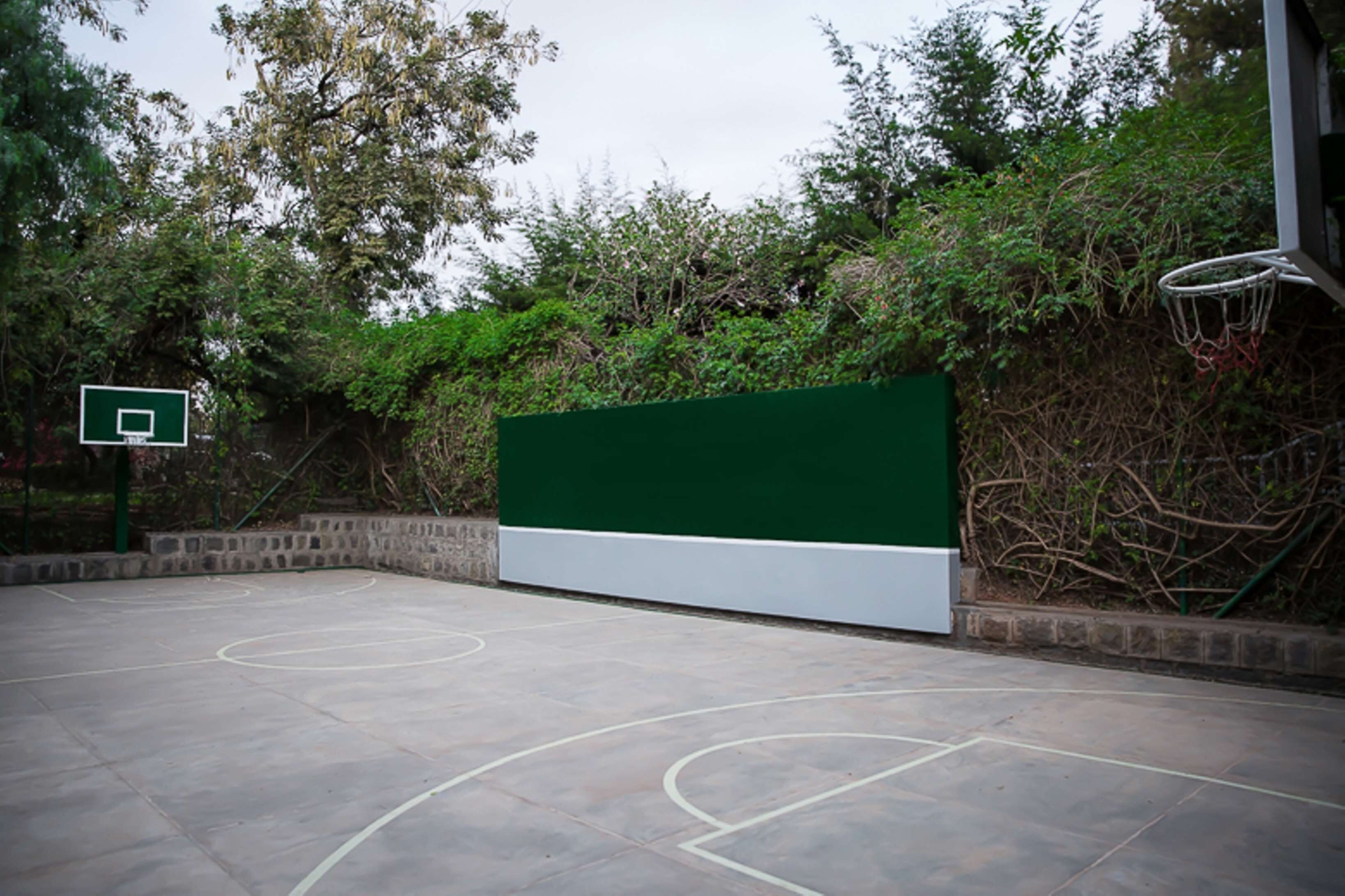Una cancha de baloncesto al aire libre con una pared verde y árboles de fondo.