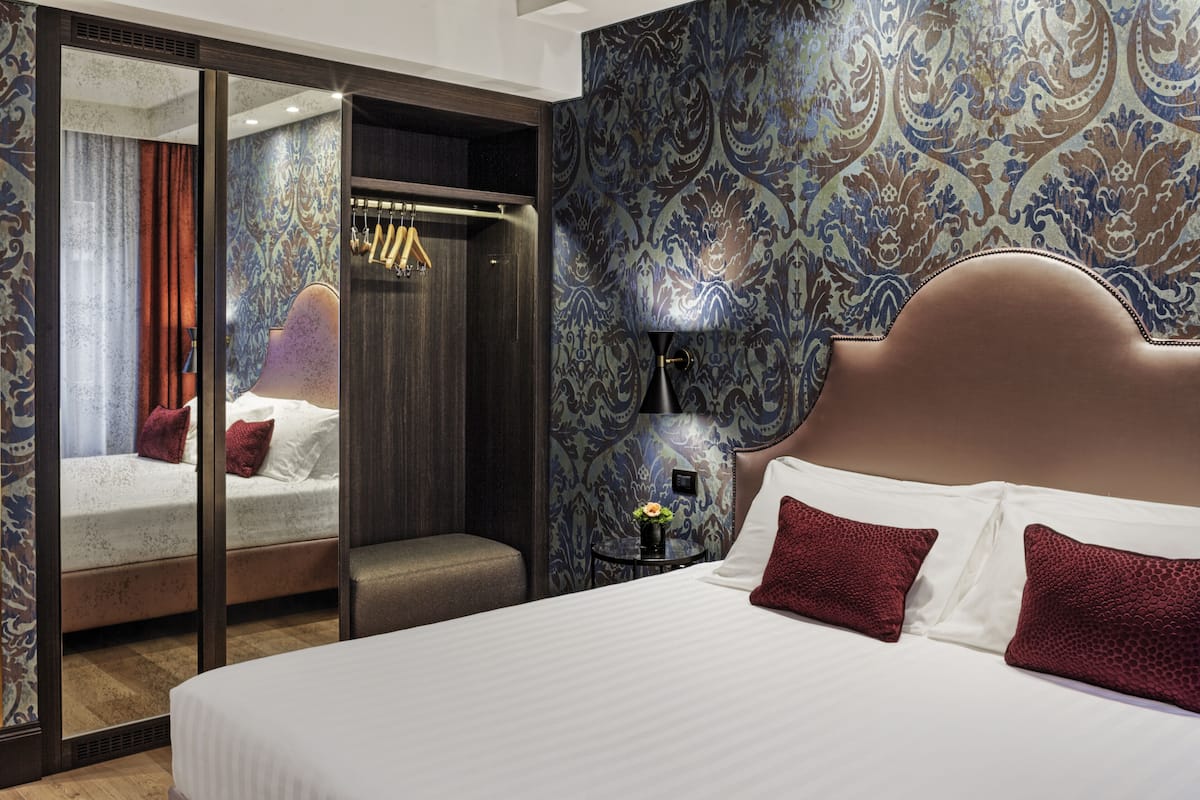 A hotel room with a large bed, a mirror, a wardrobe, and a nightstand with a lamp.