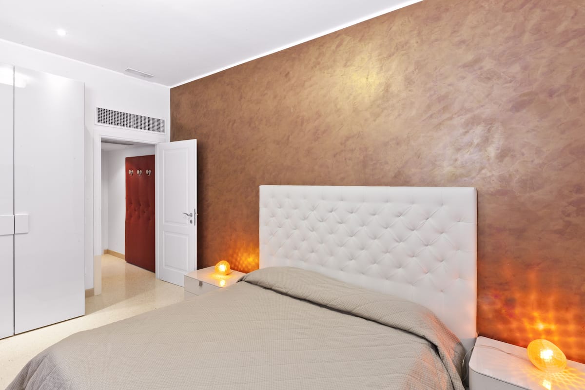 A modern bedroom with a tufted headboard, a bed with a beige quilt, bedside lamps, and a wardrobe.
