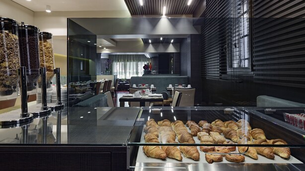 A bakery display with pastries, a seating area with tables and chairs, and a modern interior design with a sloped ceiling and glass partitions. - Hotel American Dinesen (Venice)