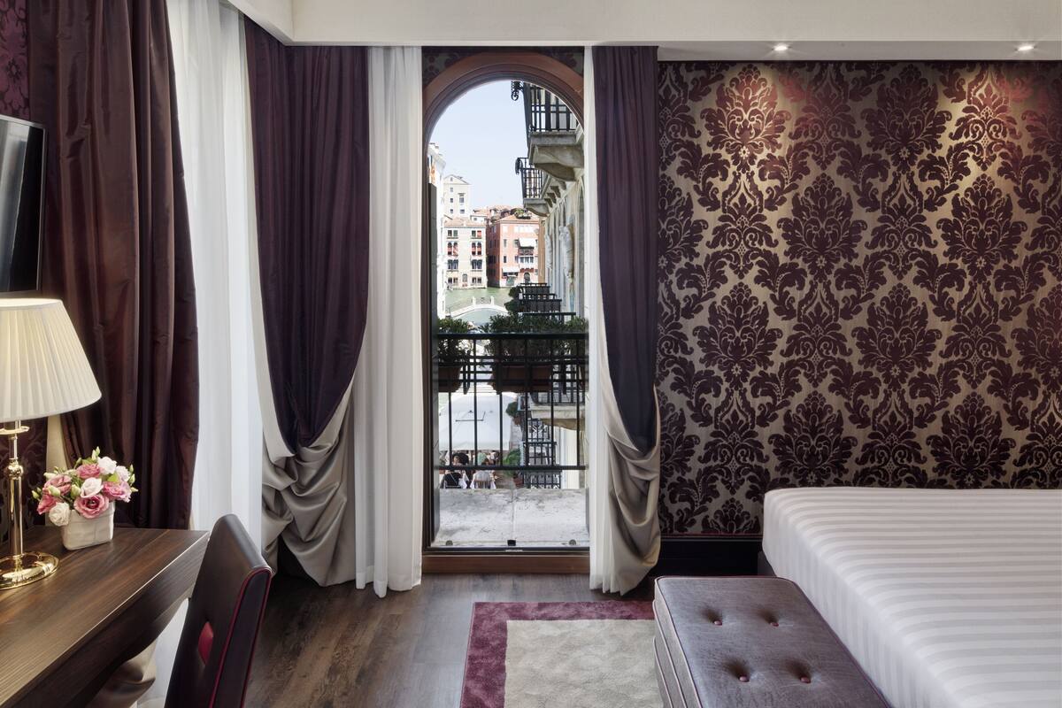 A hotel room with a bed, a desk with a lamp, a chair, a window with curtains, and a patterned wall.