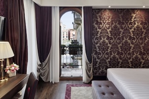 Quarto de hotel com uma cama, uma mesa com luminária, uma cadeira, uma janela com cortinas e parede estampada.