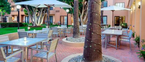 An outdoor patio area with tables and chairs, surrounded by trees and a building in the background.