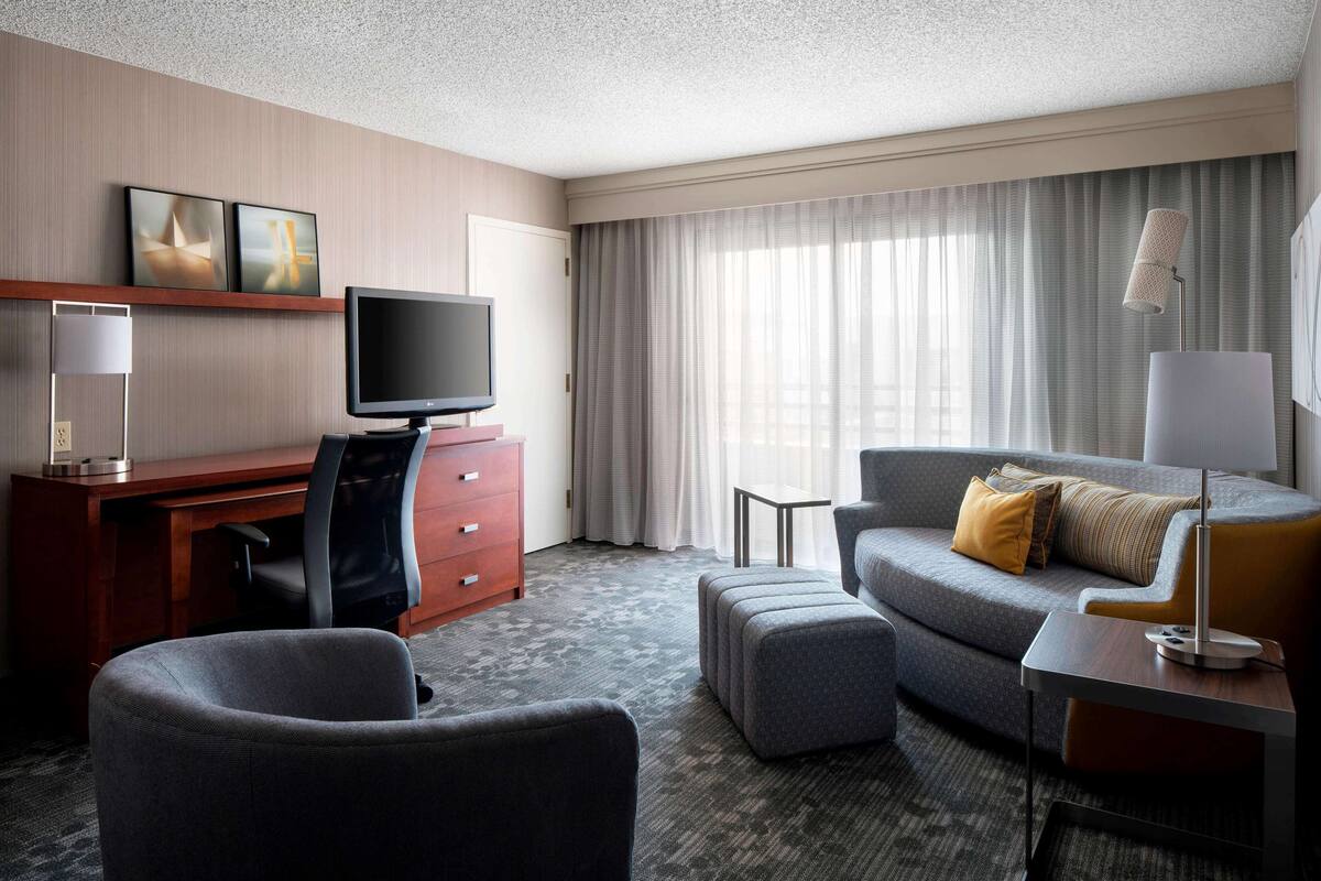 A hotel room with a flat-screen TV, a desk, a chair, a sofa, a lamp, and a window with curtains.