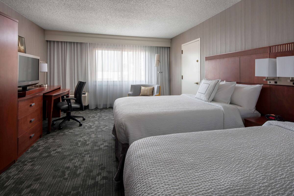 A hotel room with two beds, a desk with a TV, a chair, and a large window with curtains.