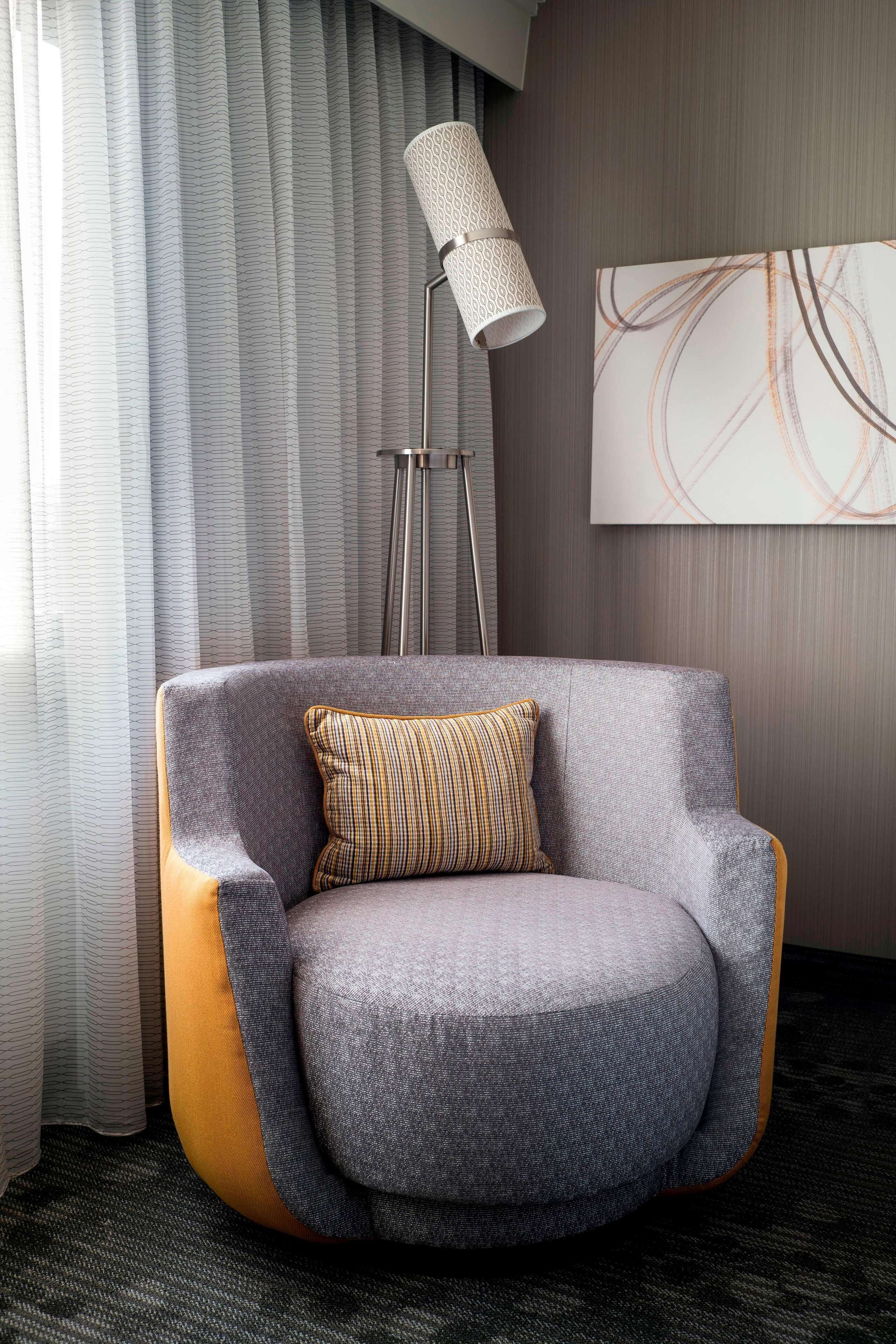 A modern armchair with a cushion, positioned in front of a floor lamp and a piece of abstract art on the wall.