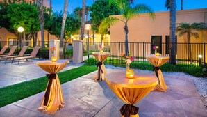 An outdoor patio area with tables draped in golden cloth, surrounded by palm trees and a building in the background.