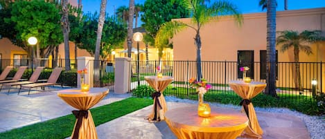 An outdoor patio area with tables draped in golden cloth, surrounded by palm trees and a building in the background.