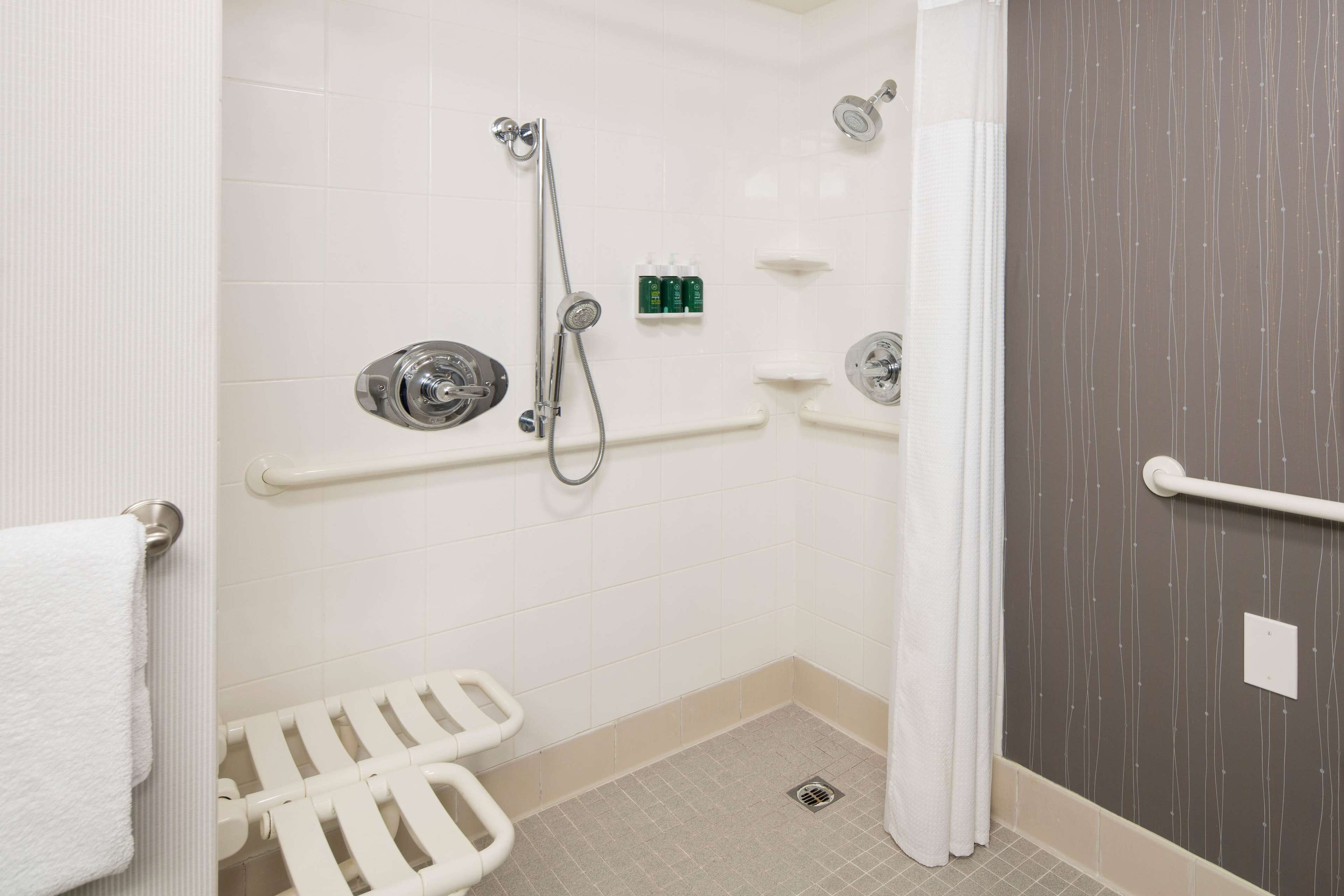 Ein Badezimmer mit weißen Fliesen, einer Dusche mit Haltegriff, einem WC mit Sicherheitsauflage und einem Handtuchhalter.