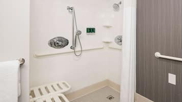 Ein Badezimmer mit weißen Fliesen, einer Dusche mit Haltegriff, einem WC mit Sicherheitsauflage und einem Handtuchhalter.