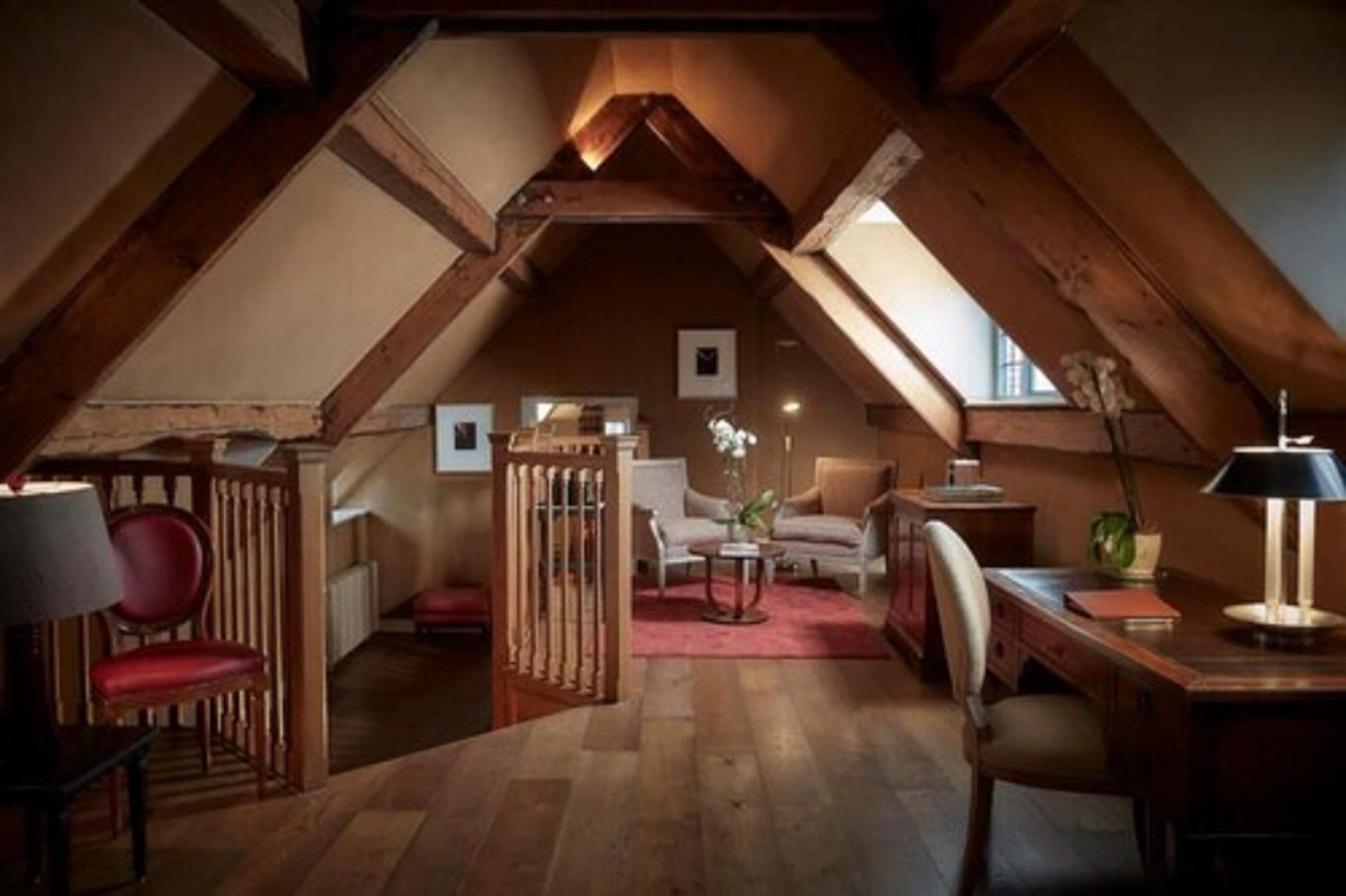 Quarto no sótão com vigas de madeira, uma escrivaninha, cadeiras e uma poltrona vermelha.
