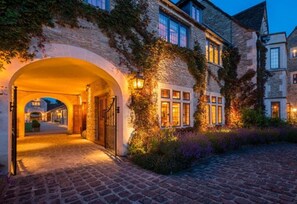 A historic stone building with an arched entrance, ivy-covered walls, and a cobblestone pathway leading to a gated driveway. - Whatley Manor (Malmesbury)