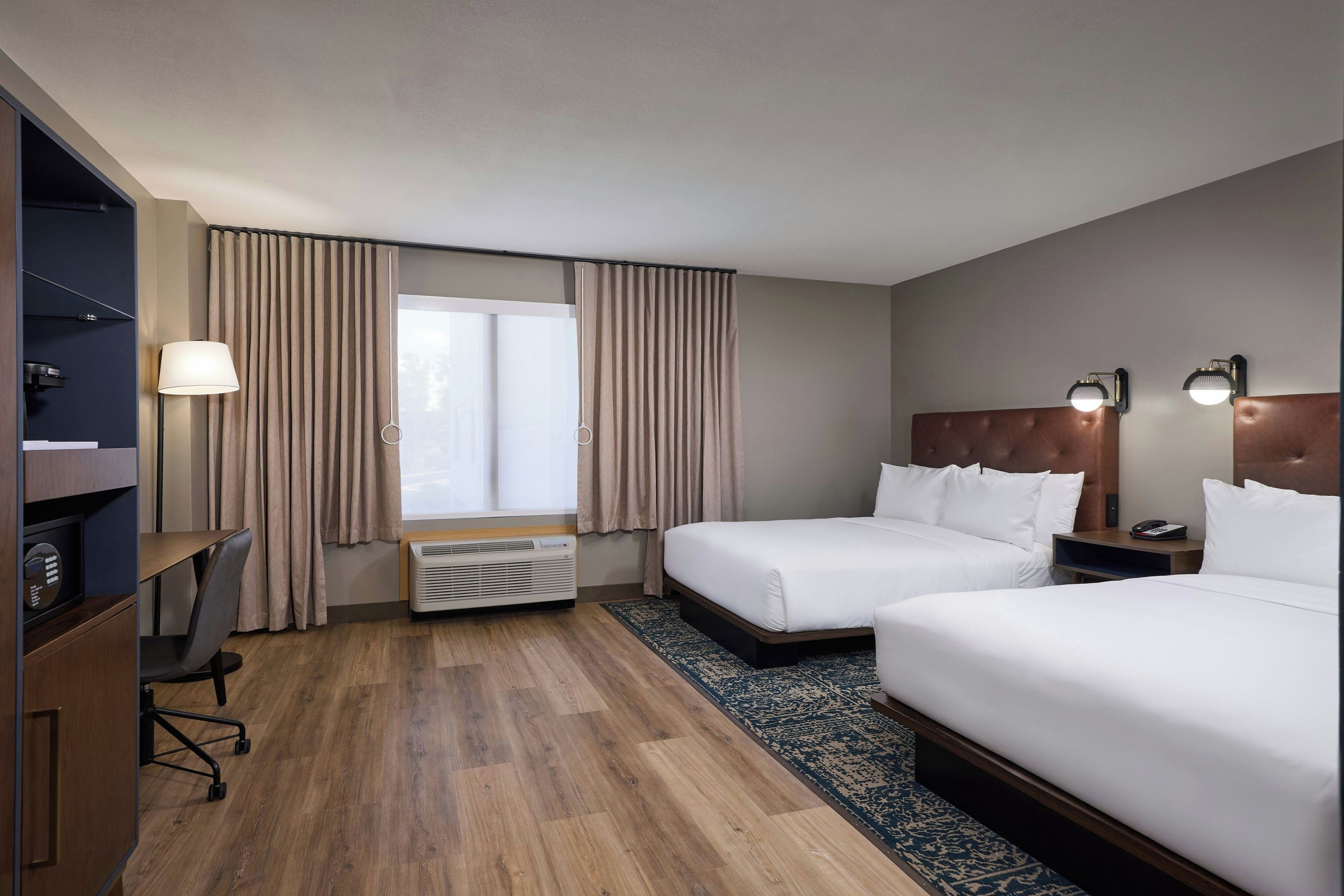 A hotel room with two beds, a desk, a chair, a heater, and a window with curtains.