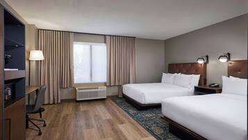 A hotel room with two beds, a desk, a chair, a heater, and a window with curtains.