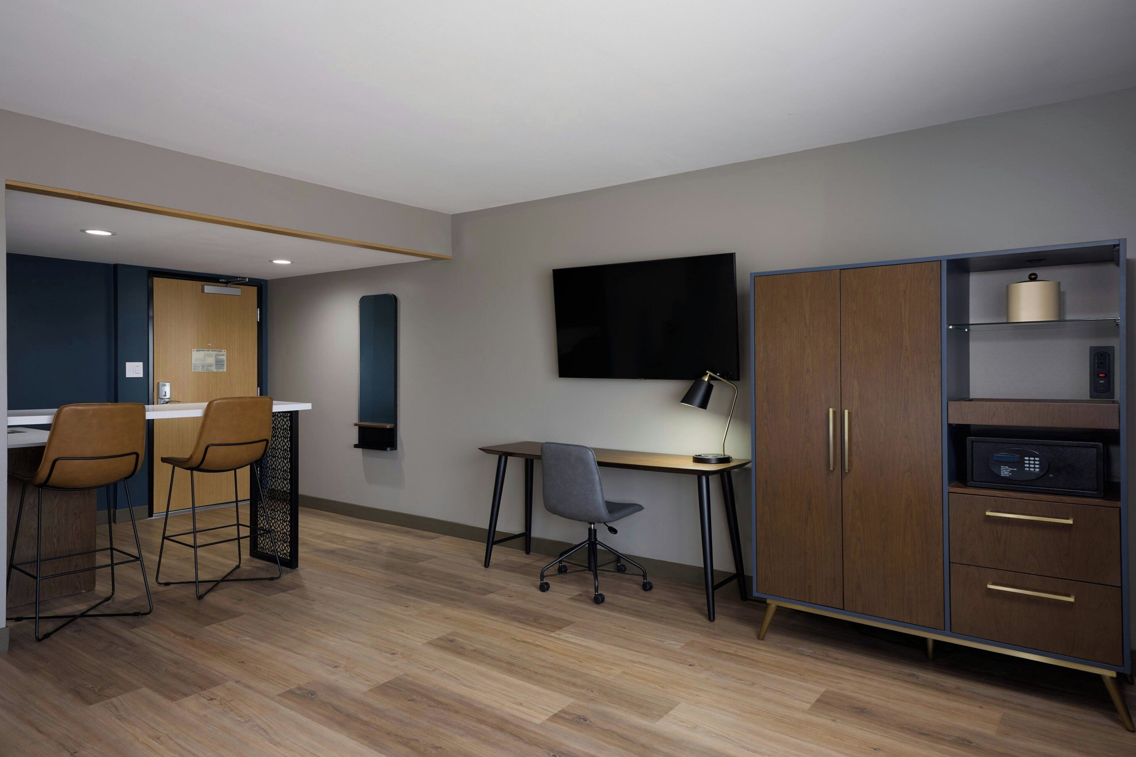 A modern office space with a desk, two bar stools, a chair, a TV, a cabinet with drawers, and a microwave.