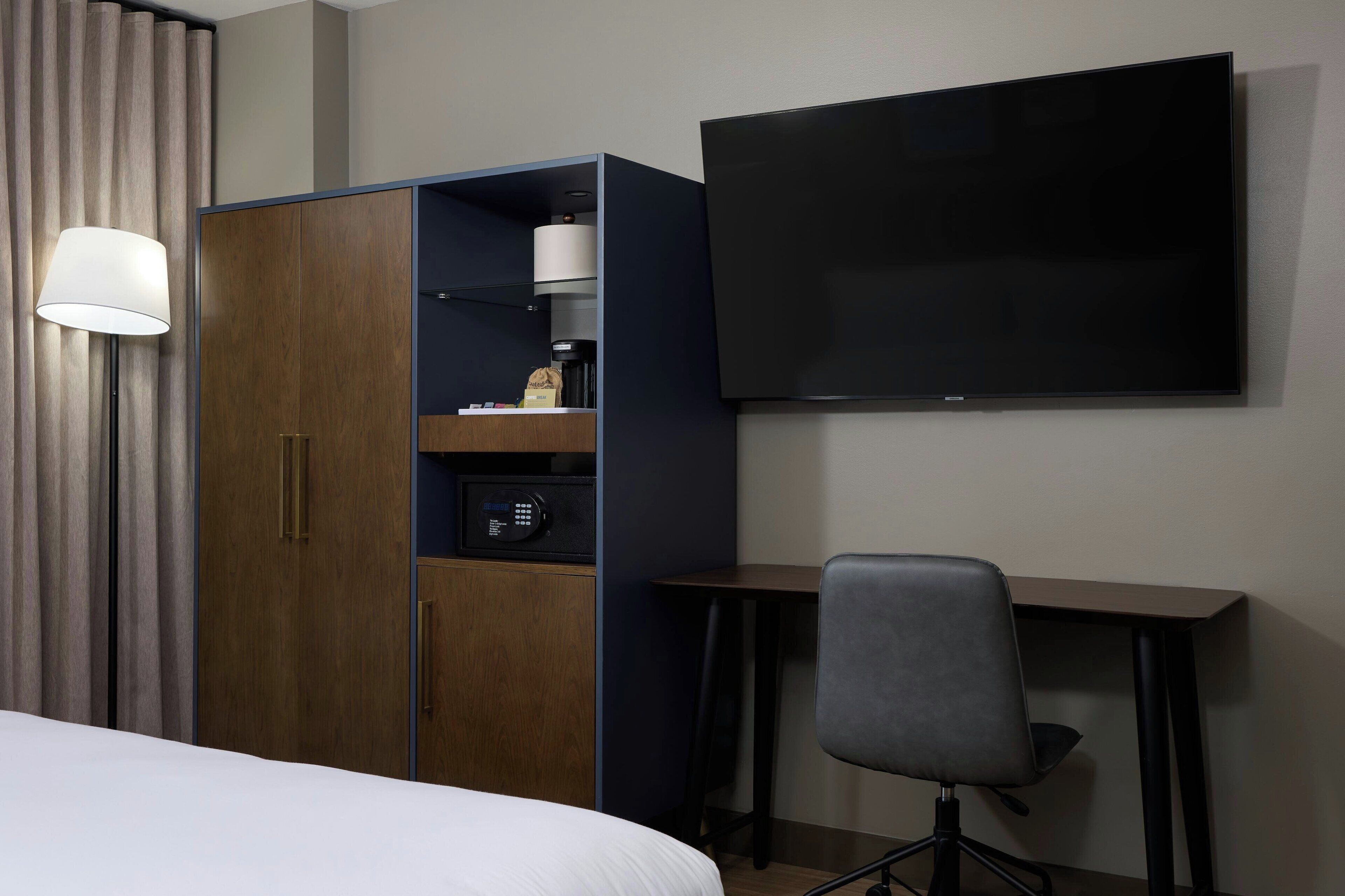A hotel room with a bed, a wardrobe, a desk, a chair, a television, and a lamp.
