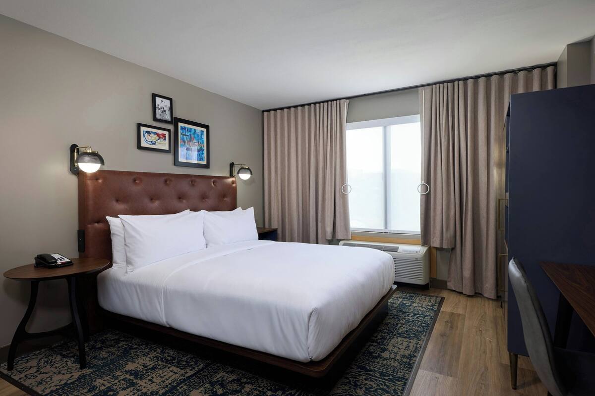A hotel room with a large bed, a bedside table with a lamp, a window with curtains, and framed pictures on the wall.