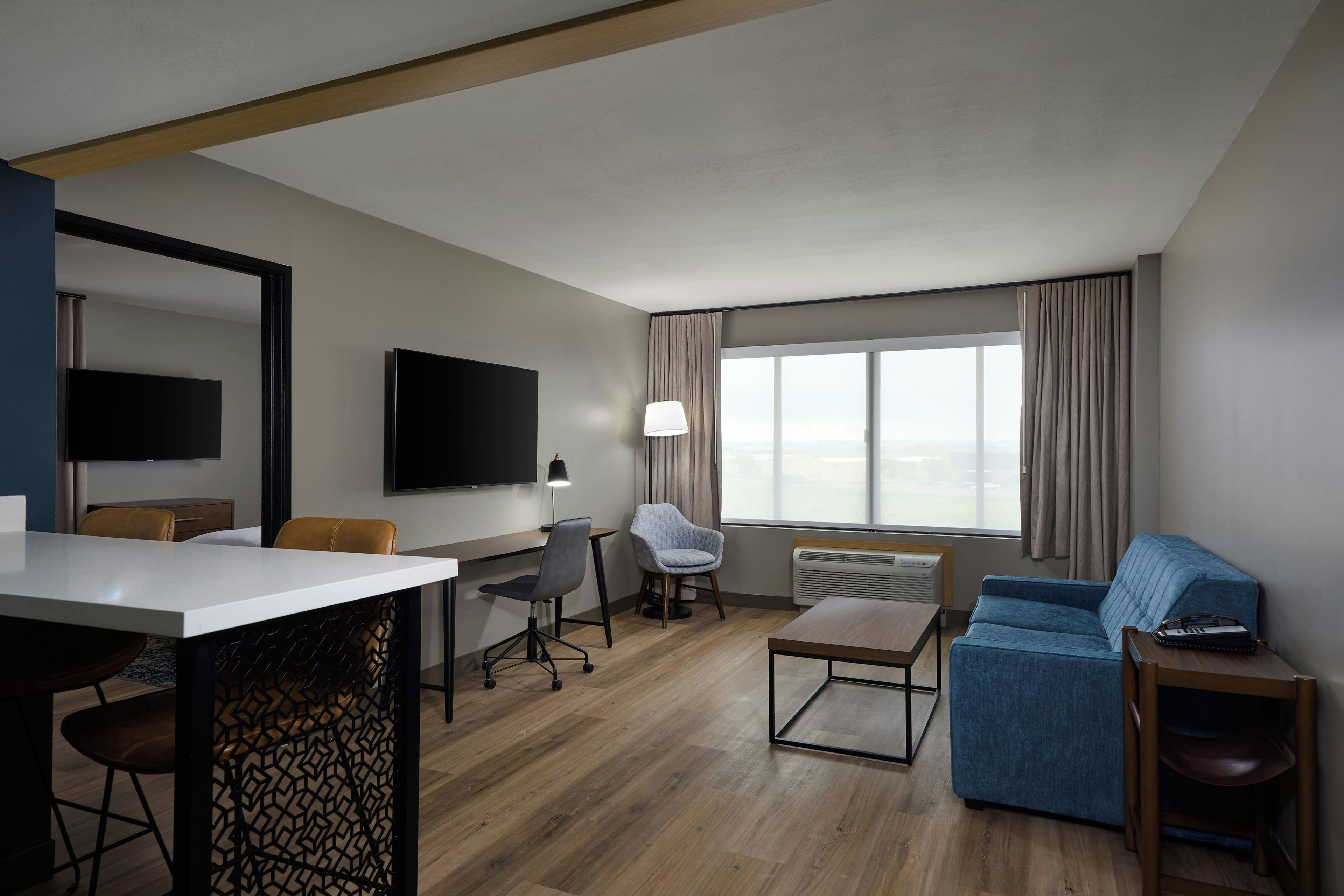 A modern hotel room with a flat-screen TV, a desk with a chair, a sofa, and a large window with curtains.