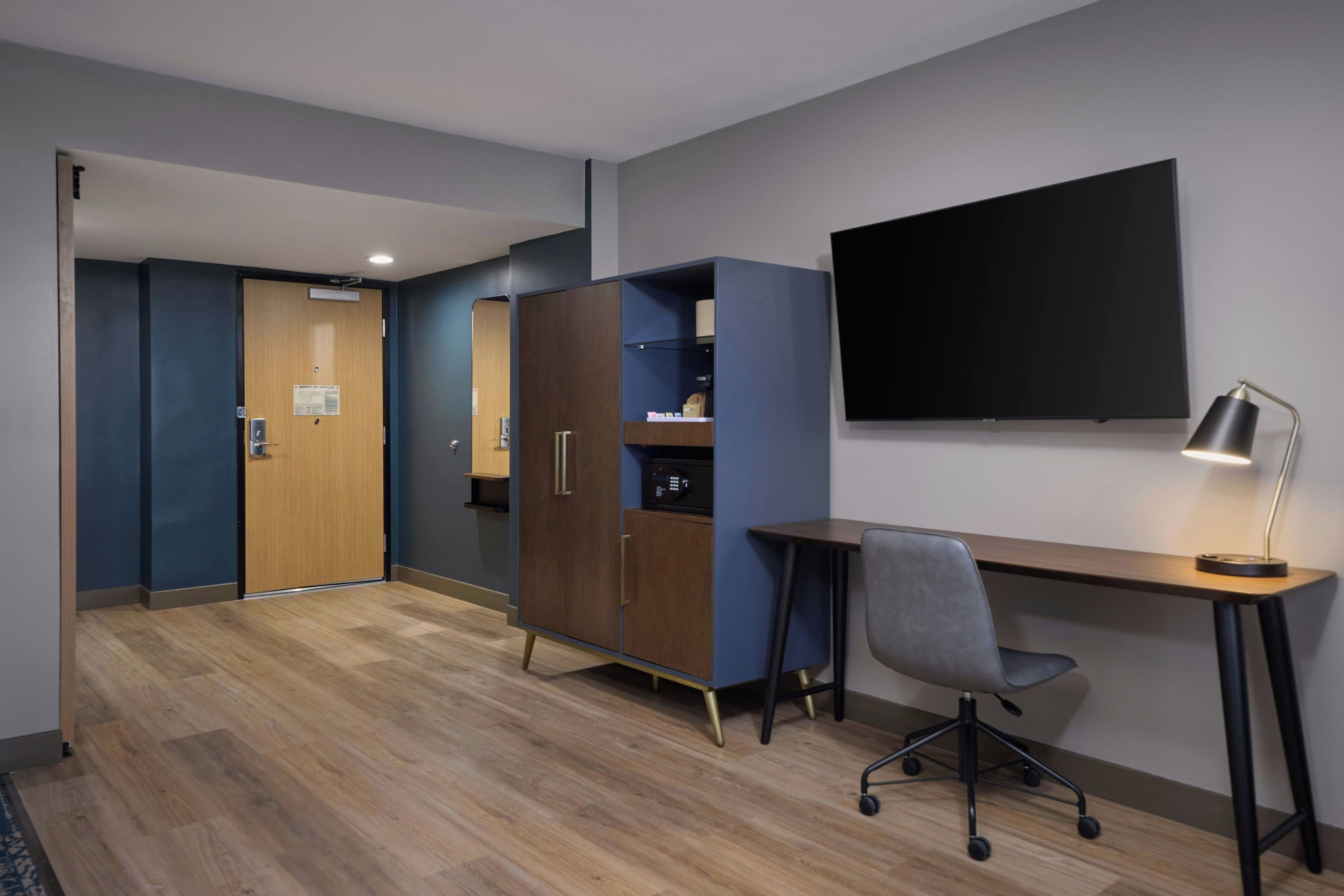 A hotel room with a wooden desk, a chair, a television mounted on the wall, and a wardrobe.
