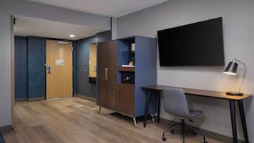 A hotel room with a wooden desk, a chair, a television mounted on the wall, and a wardrobe.