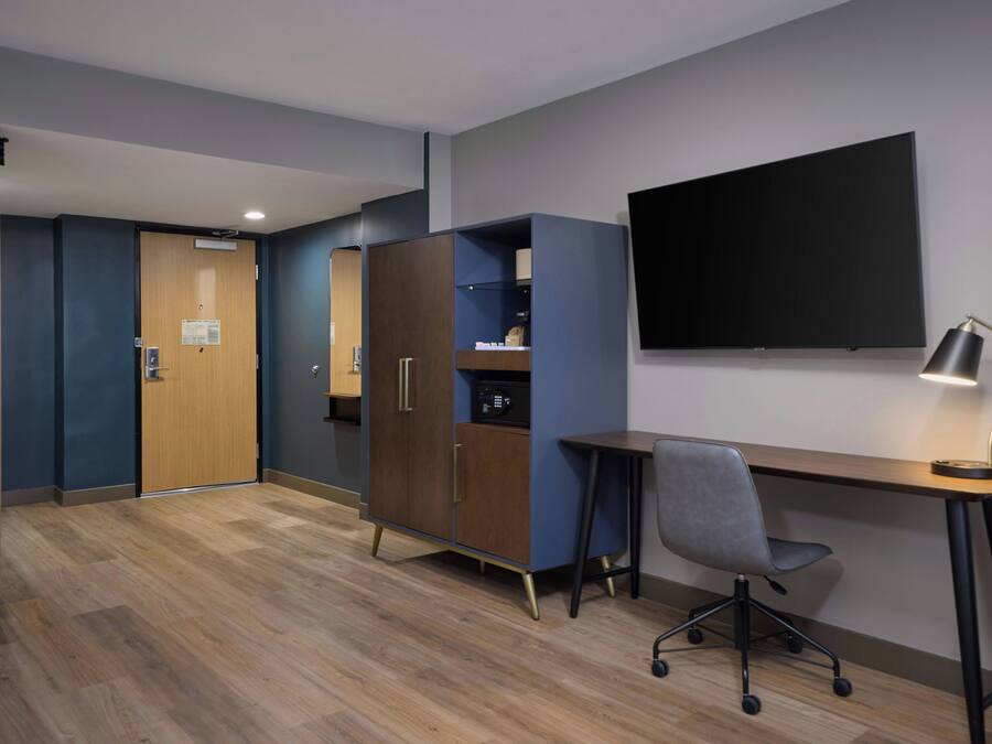 A hotel room with a wooden desk, a chair, a television mounted on the wall, and a wardrobe.