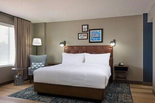 Habitación de hotel con una cama grande, una silla, una mesita de noche, una lámpara y una obra de arte en la pared.