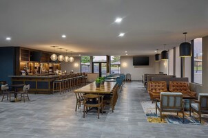 Uma sala de jantar ampla com uma mesa de madeira comprida e várias cadeiras, uma área de bar com balcão e banquetas, e um espaço de estar com sofás e cadeiras.