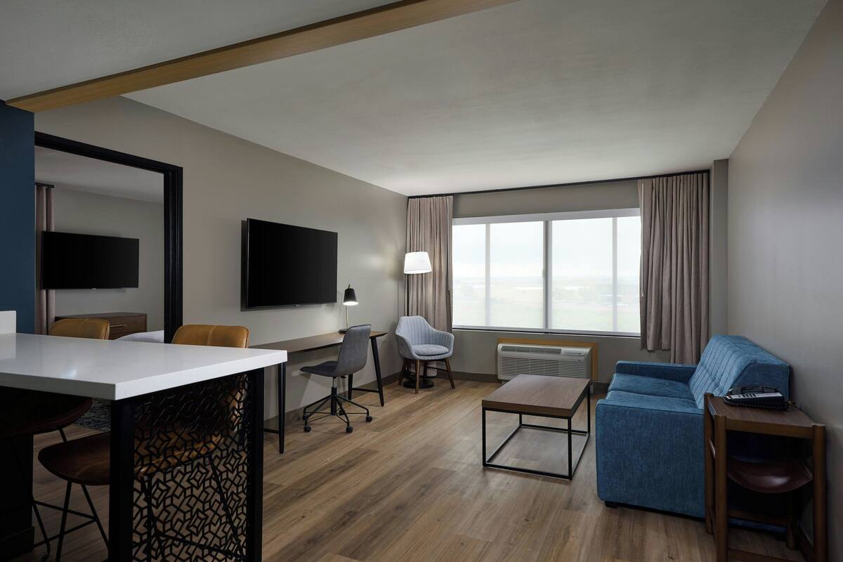 A modern hotel room with a wooden floor, a flat-screen TV mounted on the wall, a desk with a chair, a sofa, and a small table with a lamp.
