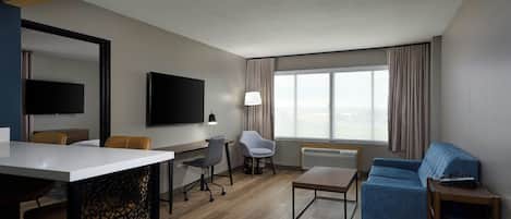 A modern hotel room with a wooden floor, a flat-screen TV mounted on the wall, a desk with a chair, a sofa, and a small table with a lamp.