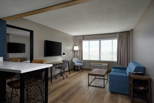Una habitación de hotel moderna con suelo de madera, un televisor de pantalla plana montado en la pared, un escritorio con silla, un sofá y una mesita de noche con lámpara.