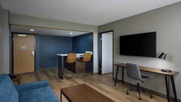 Una habitación de hotel moderna con un sofá azul, un escritorio de madera, dos sillas, un televisor y una puerta que da a una zona de cocina.