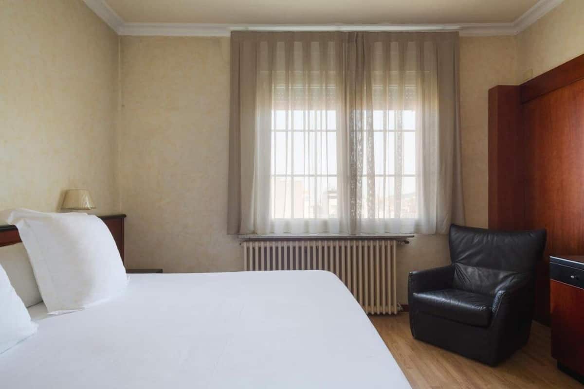 Een hotelkamer met een groot bed, een zwarte leren fauteuil, een houten garderobe en een raam met dunne gordijnen.