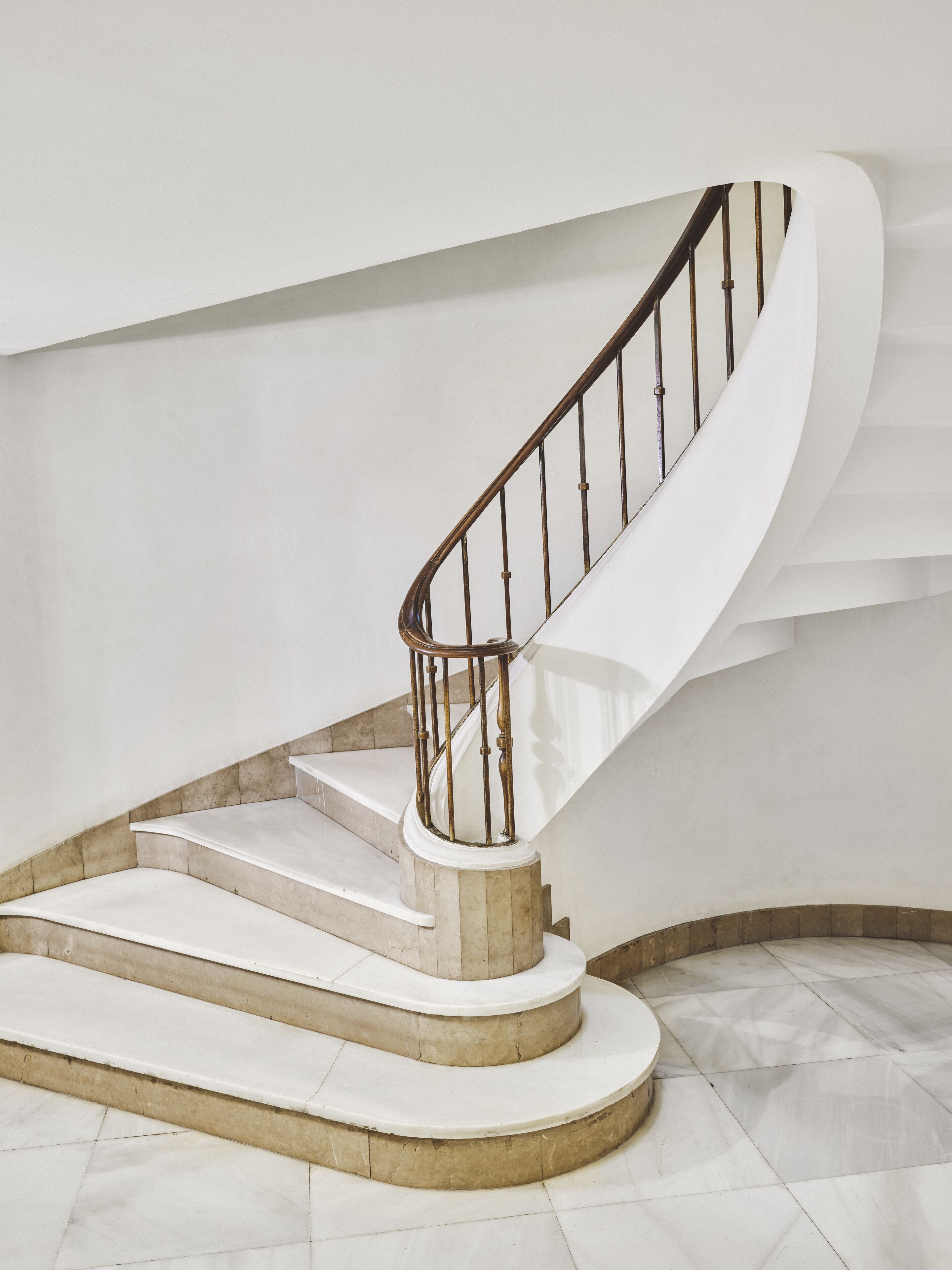Una escalera curva con barandilla de metal, escalones de mármol y pared blanca.