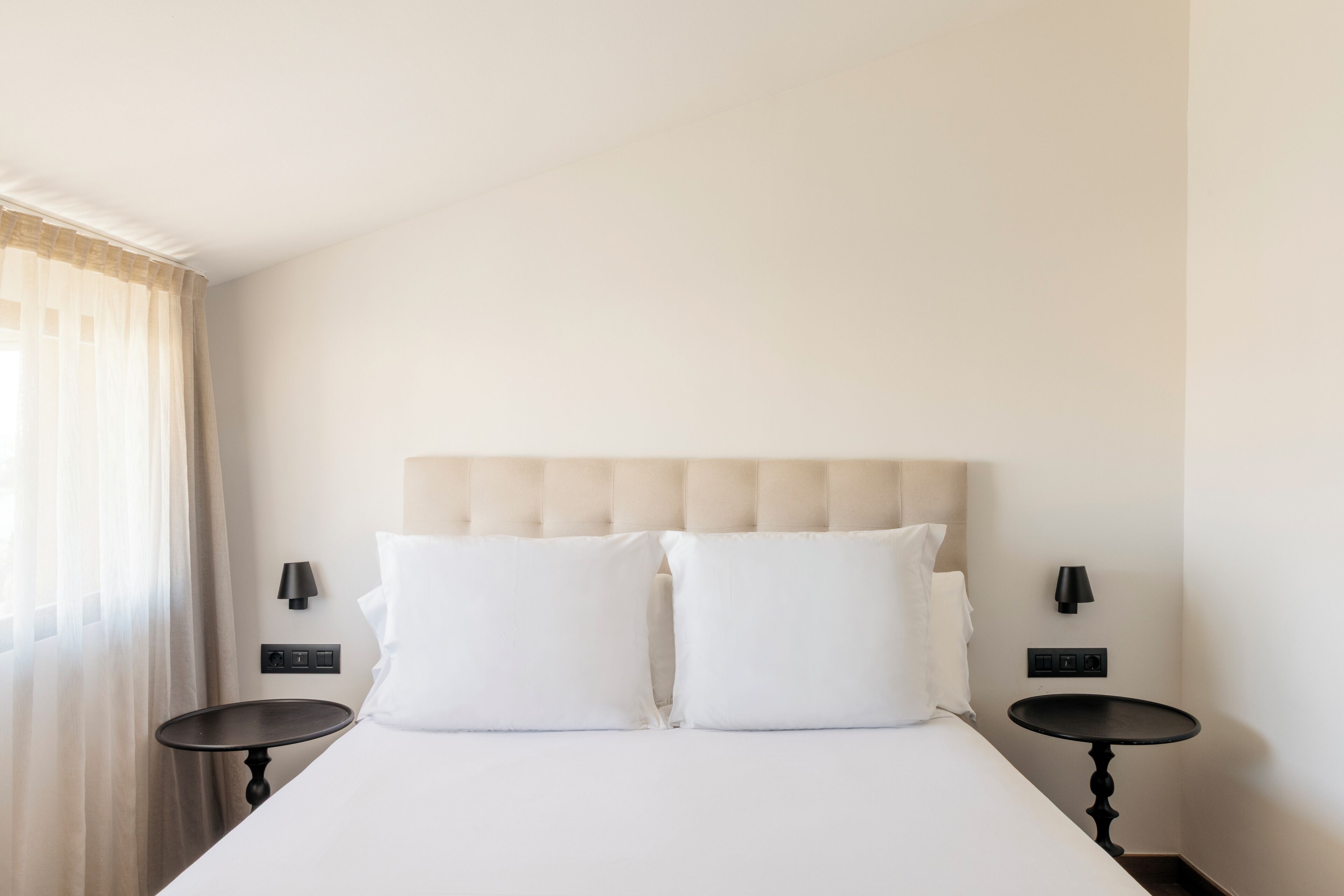 Una habitación de hotel con una cama, dos mesitas de noche, un cabecero y una ventana con cortinas.
