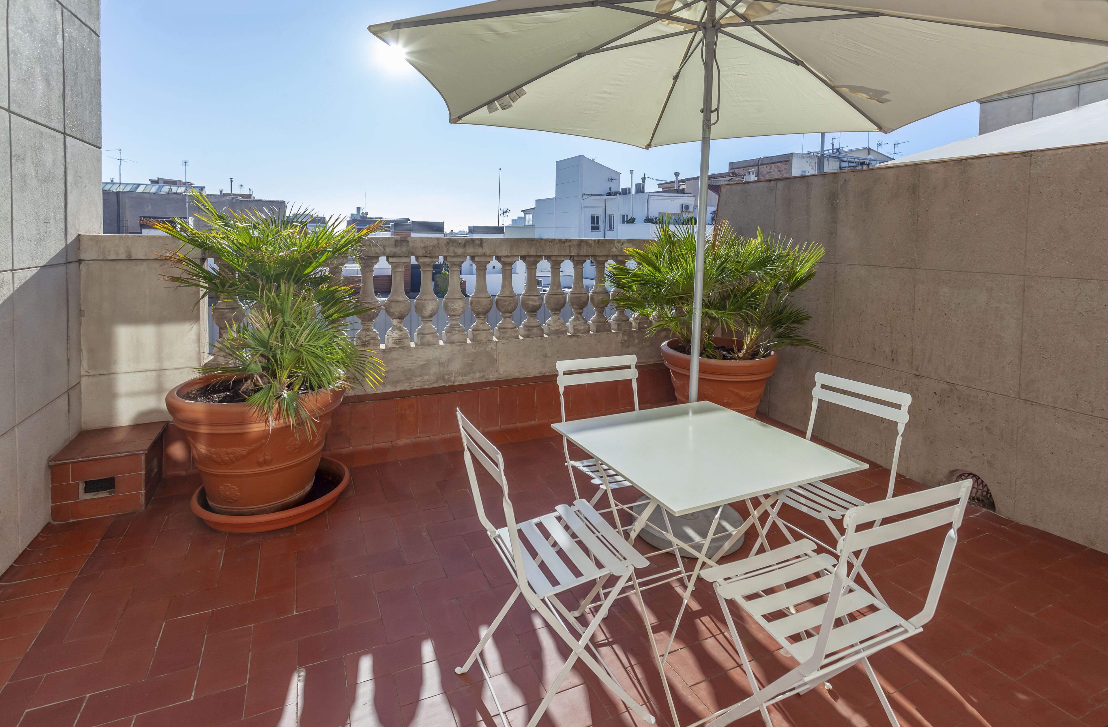 Terrassa al terrat amb taula i cadires blanques, dues plantes en testos i un parasol gran.
