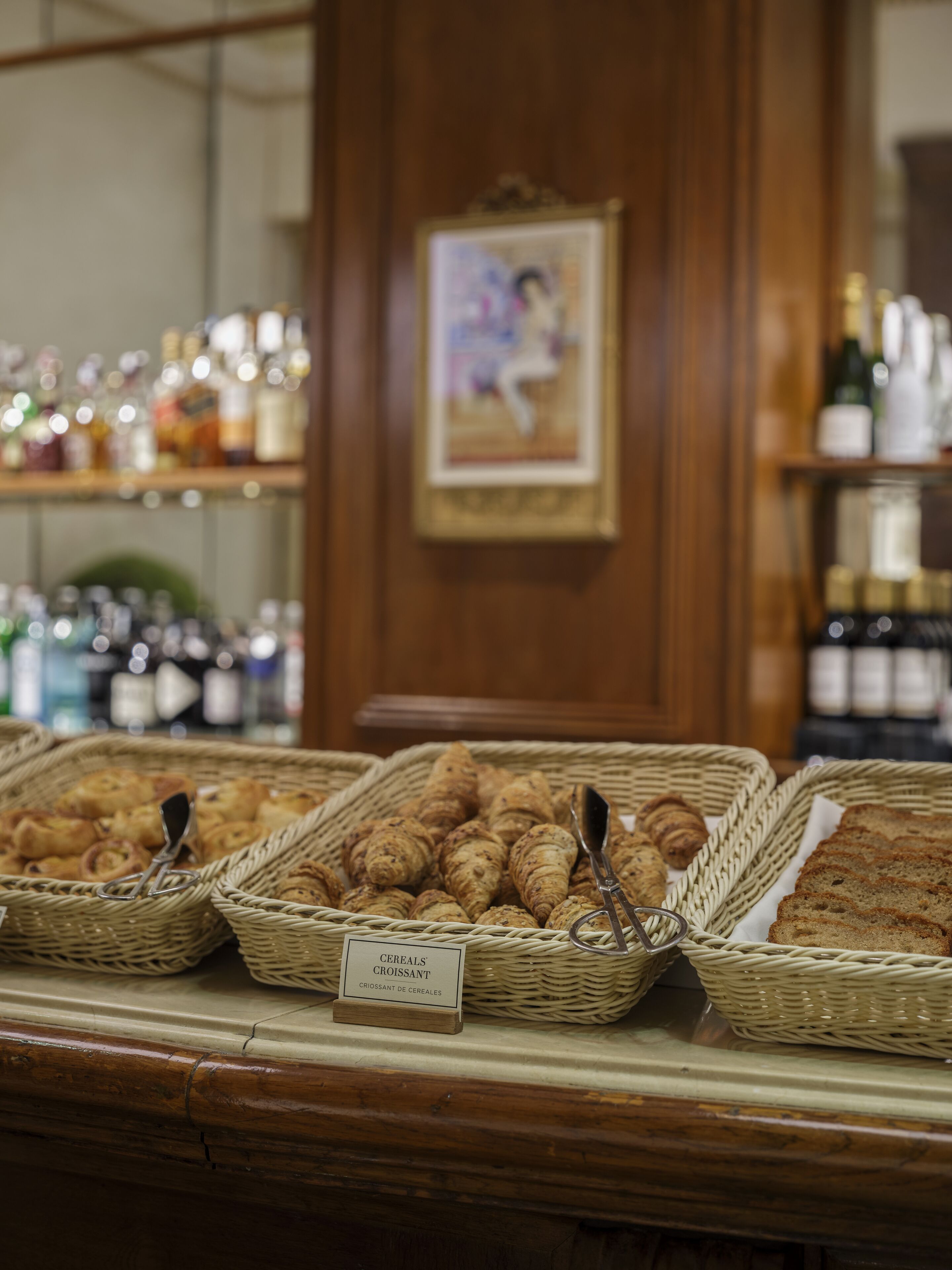 Une vitrine de boulangerie avec des paniers remplis de viennoiseries et de pain, une enseigne indiquant « CEREALS’ CROISSANT », et des étagères avec des bouteilles en arrière-plan.