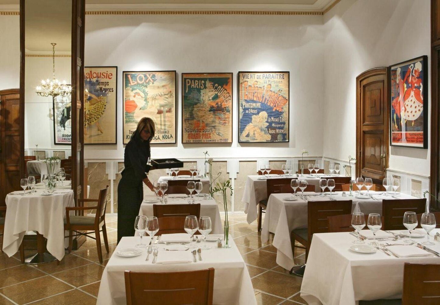 Un restaurant avec plusieurs tables dressées pour le service, un serveur s’occupant d’une table et des affiches accrochées au mur.