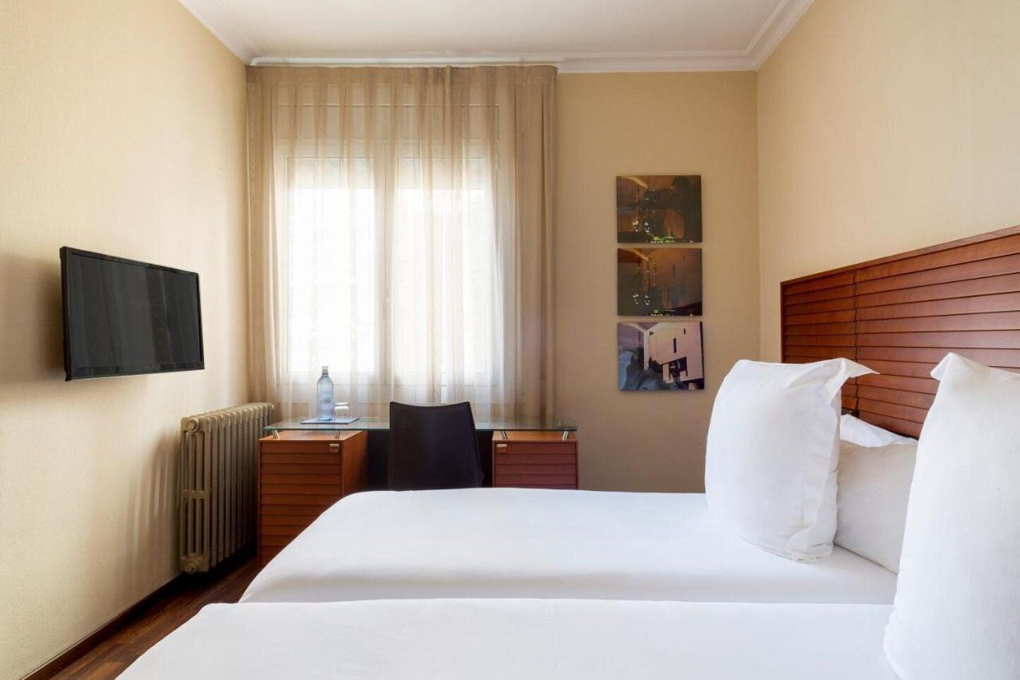 A hotel room with two beds, a desk, a chair, a radiator, a television, and a window with curtains.