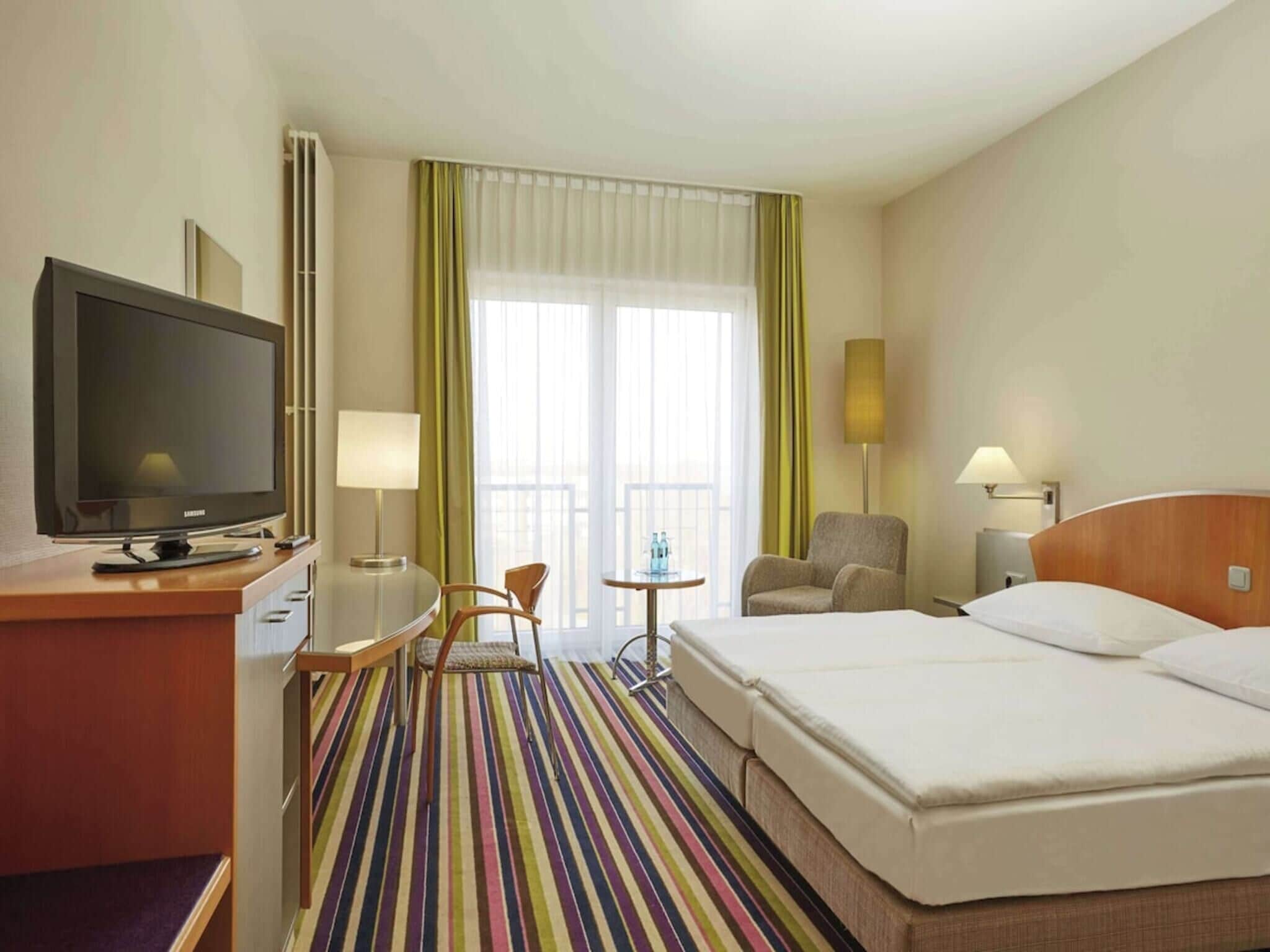 A hotel room with a bed, a desk with a television, two chairs, a small table, and a window with curtains.