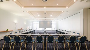 Un salón de conferencias con una larga mesa, sillas, una pantalla de proyector y piso alfombrado.