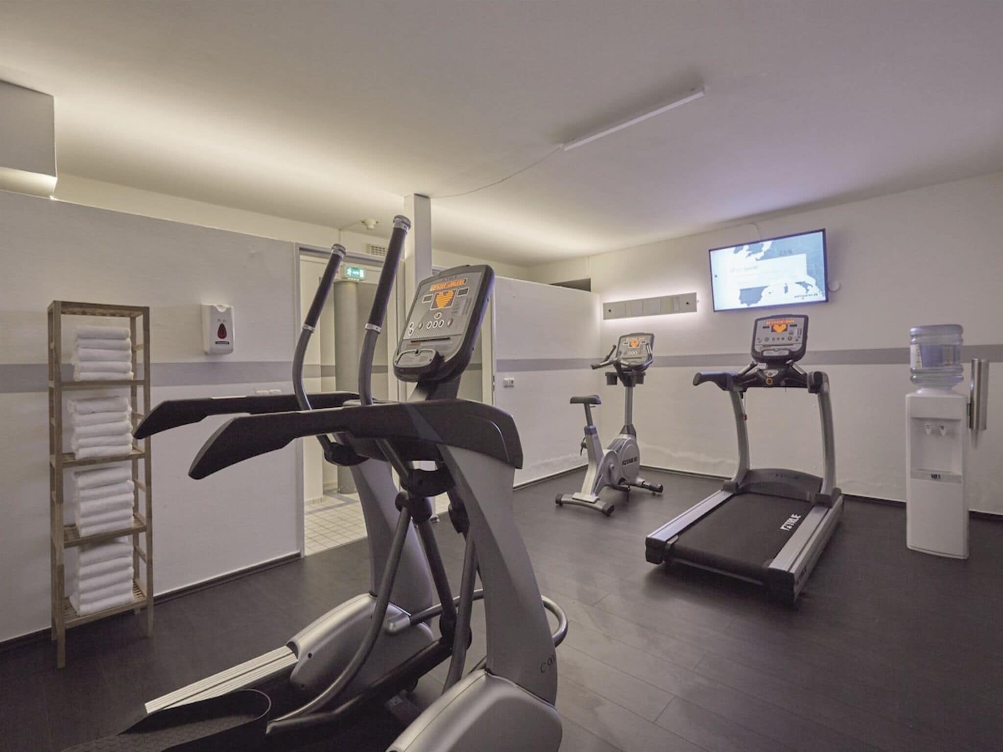 Centro de fitness com esteiras, bicicleta ergométrica e tela na parede.