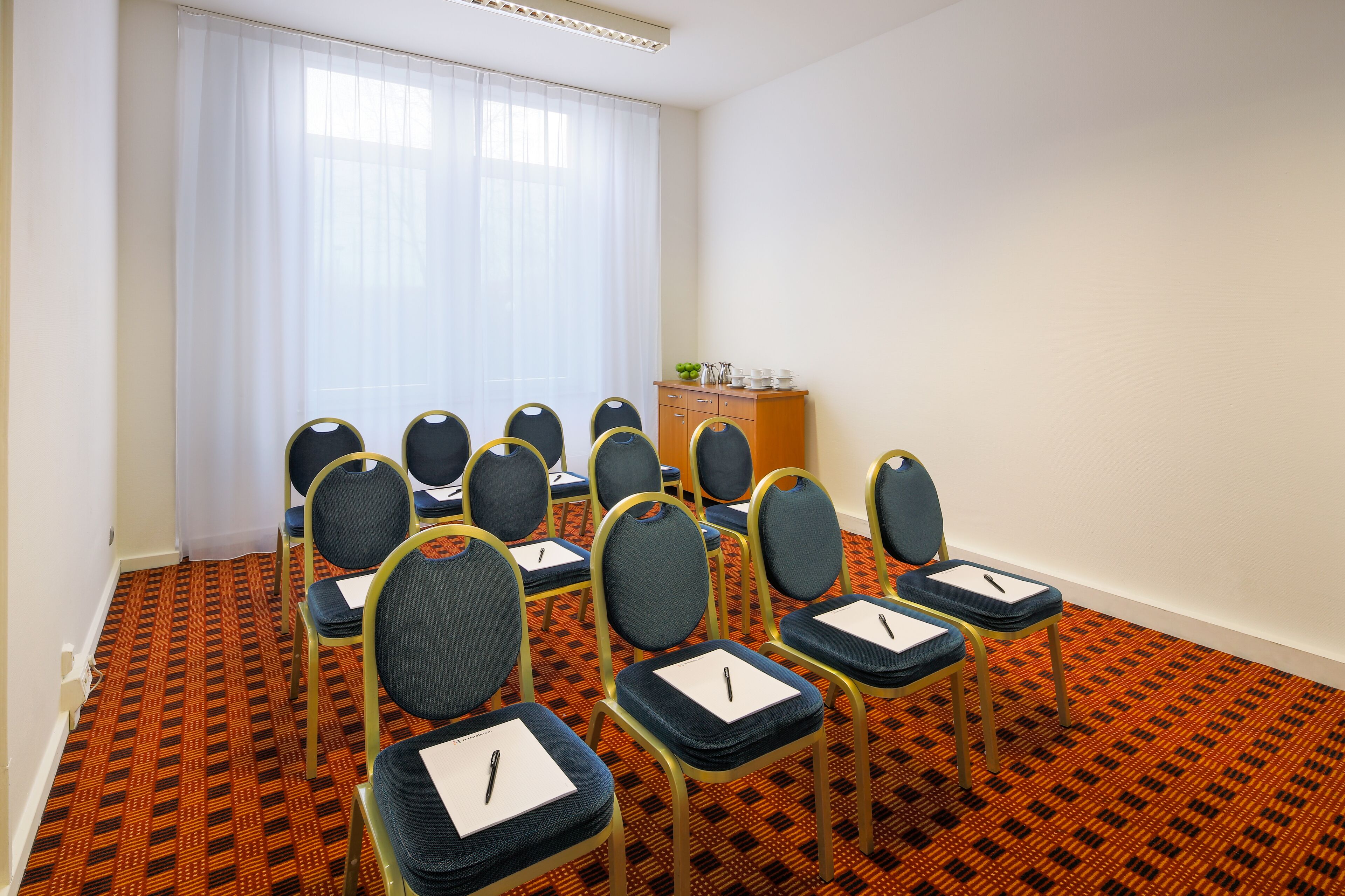 A conference room with rows of chairs, a wooden cabinet with items on top, and a patterned carpet.