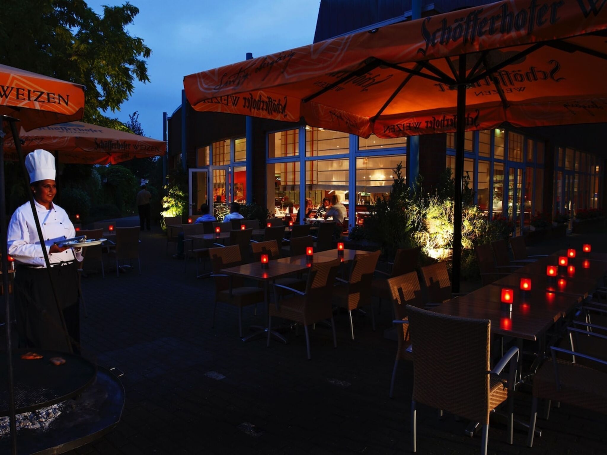 An outdoor dining area with tables and chairs, a chef standing by a grill, and a building with large windows in the background.